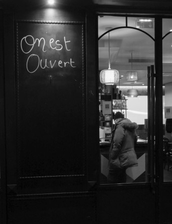 Une façade de café avec "On est ouvert" écrit dessus et un homme seul au comptoir. Jeu de mot sur l'idée de couple ouvert et mis en avant de la solitude.