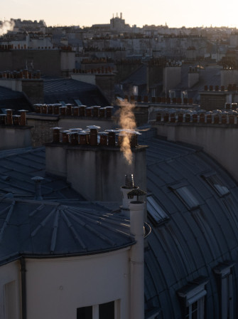 De la fumée, teintée par le soleil doré du matin, s'élève d'une cheminée parisienne.