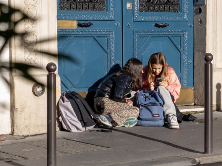 Deux jeunes filles se parlent avec animation, tôt le matin, assises devant la porte bleue d'un immeuble parisien, rue de Clichy.